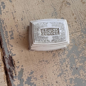 May include: A rectangular, white jewelry box with a floral design. The box has a hinged lid and is decorated with intricate floral patterns and a central rectangular panel. The box is made of a light-colored material and has a silver trim.