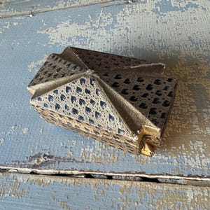 May include: A vintage, geometric-shaped box with a gold-colored exterior and a patterned top featuring a repeating dark blue design. The box is resting on a distressed, light blue wooden surface.