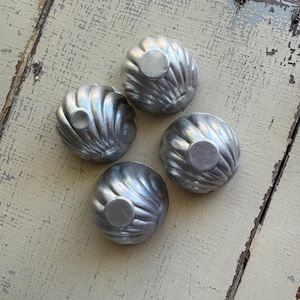 May include: Four vintage, silver-colored metal molds with a fluted design. Each mold has a circular, raised center. The molds are arranged on a distressed, light-colored wooden surface, creating a rustic aesthetic. These are likely used for baking or creating decorative food items.