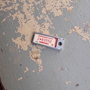May include: A small, rectangular Nebraska license plate with the text "Cornhusker State" and the numbers "2-B3552" in red. The year "1974" and the number "72" are also visible. The plate is set against a weathered, gray and beige surface.