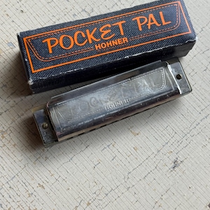 May include: A silver harmonica with the text "POCKET PAL HOHNER" is displayed next to its black box. The box has orange text and trim. The harmonica is on a distressed, white-painted wooden surface.