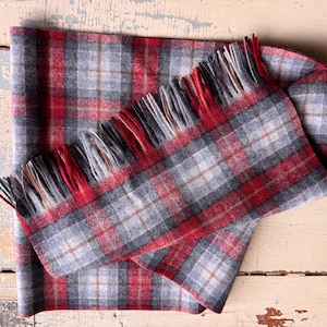May include: A rectangular scarf featuring a plaid pattern in shades of red, grey, and white with thin brown accent lines. The scarf has a soft, felted texture and is finished with short, fringed edges along the width, displaying alternating colors.