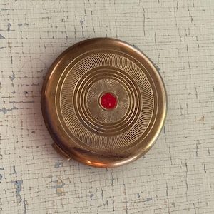 May include: A round, metallic gold-toned compact case featuring a series of concentric circular grooves etched into the surface. A small, circular, vibrant red accent is positioned directly in the center of the lid, providing a distinct contrast.