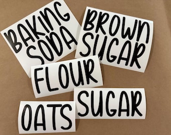 May include: White vinyl labels with black lettering for kitchen pantry organization. The labels read "BAKING SODA", "BROWN SUGAR", "FLOUR", "OATS", and "SUGAR". The labels are arranged on a brown surface.