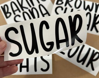 May include: Close-up of white labels with black text for kitchen organization. The word "SUGAR" is prominently displayed on one label, with other labels reading "BAKING SODA", "BROWN SUGAR", "FLOUR", and "THIS".