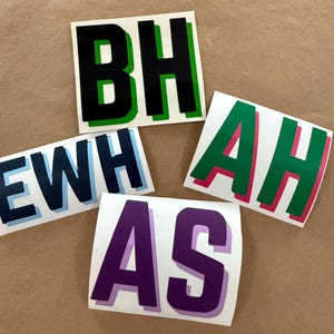 May include: Four square vinyl decals featuring bold block letters. One decal displays BH in black with a green shadow, EWH in dark blue with a light blue shadow, AH in green with a pink shadow, and AS in purple with a light purple shadow.