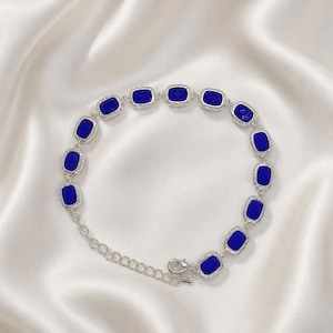 May include: A silver-toned bracelet featuring a series of rectangular, deep blue faceted stones set in metallic frames. The bracelet includes a delicate chain link section and a lobster claw clasp, creating a continuous circular arrangement.