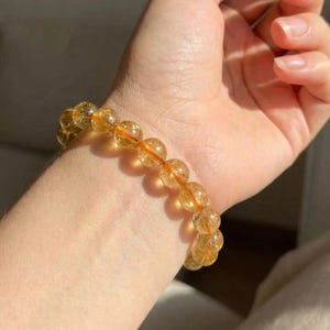 Op de afbeelding: Een armband met doorschijnende, goudgele citrien edelsteen kralen. De armband is geregen op een dun, oranje elastisch koord. De armband wordt om een pols gedragen, met een zilveren ring om een vinger.