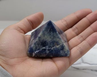 May include: A small, pyramid-shaped stone with a dark blue and green mottled pattern. The crystal is held in an open hand, showcasing its geometric form and natural colour variations. The stone is likely a decorative item or a healing crystal.