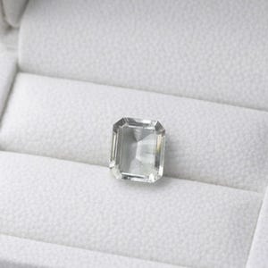 May include: A rectangular, pale green gemstone with a faceted cut, presented in an open, white, velvet-lined jewellery box. The box highlights the gem's clarity. The setting is simple, focusing on the stone's natural beauty and sparkle.