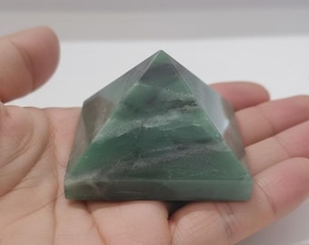 May include: A green and gray pyramid-shaped crystal. The pyramid is held in a hand, showcasing its geometric form and natural color variations. The crystal appears to be polished, with smooth surfaces and a translucent quality.