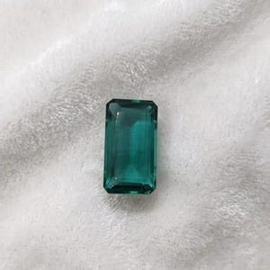 May include: A rectangular, emerald-cut gemstone in a deep teal hue. The gem is displayed against a soft, white backdrop, accentuating its clarity and facets. The gemstone's form is a classic rectangle with cut corners.