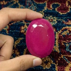 以下が含まれることがあります： ファセット加工された、楕円形のマゼンタ色の宝石。 宝石は指の間に挟まれ、研磨された表面と、多くのファセットに光が当たる様子を示しています。 背景は、青とブルゴーニュの色合いの模様の入ったラグです。