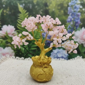 Puede incluir: Un árbol decorativo de cristal de cuarzo rosa con una base dorada en forma de bolsa de dinero. Las ramas del árbol son de alambre dorado y las hojas son pequeños cristales de color rosa claro. El fondo está borroso con flores.