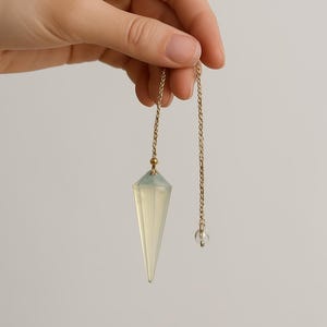 Puede incluir: Un péndulo con una cadena dorada y un cristal transparente y puntiagudo. El cristal es sostenido por una mano contra un fondo liso. El péndulo es una herramienta utilizada para la adivinación y las prácticas espirituales.