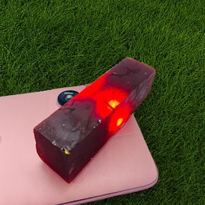 Könnte beinhalten: Ein rechteckiger, dunkelroter Gegenstand mit leuchtend rotem Inneren, der auf einer rosa Oberfläche ruht. Der Gegenstand hat ein raues, strukturiertes Aussehen und scheint aus einem durchscheinenden Material zu bestehen. Der Hintergrund ist grünes Gras.