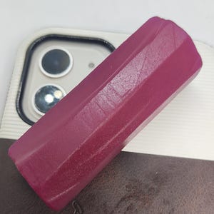 Op de afbeelding: Een gefacetteerd, rechthoekig object in diep magenta. Het object heeft een getextureerd oppervlak en lijkt van een vast materiaal te zijn gemaakt. Het object ligt op een wit en bruin oppervlak, mogelijk een telefoonhoesje.