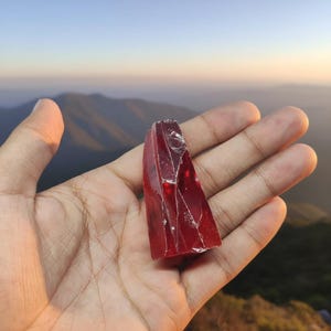 Op de afbeelding: Een doorschijnend, robijnrood, driehoekig kristal met interne breuken. Het kristal wordt in een open hand gehouden tegen een achtergrond van een bergketen en een gradiënthemel.