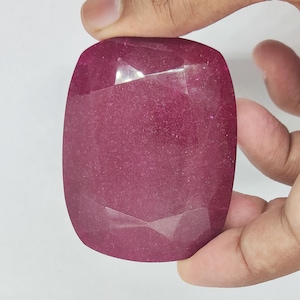 May include: A large, faceted, cushion-cut ruby gemstone. The ruby is a deep, rich red color with visible internal inclusions. The gemstone is held between two fingers against a white background. The cut of the stone is designed to maximize brilliance.