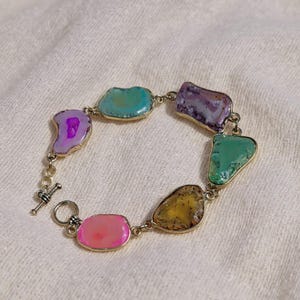 May include: A gemstone bracelet with polished stones in pink, purple, green, yellow, and blue. The stones are set in gold-coloured metal and linked by a delicate chain. The bracelet has a toggle clasp. The bracelet is approximately 18 cm in length.