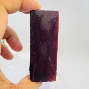 Puede incluir: Una gema rectangular y en bruto de color burdeos intenso. La piedra tiene una superficie rugosa y texturizada y se sostiene en una mano sobre un fondo blanco. La gema parece ser un espécimen mineral natural.