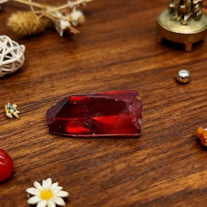 Op de afbeelding: Een gefacetteerde, dieprode edelsteen met een ruwe rand, rustend op een houten oppervlak. De edelsteen heeft een rijke, transparante rode kleur. De afbeelding bevat ook kleine decoratieve items, zoals een witte bal en een klein messing object.