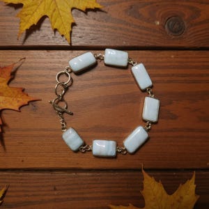 May include: A bracelet featuring rectangular, light blue gemstones set in gold-colored metal. The bracelet has a toggle clasp and is displayed on a wooden surface with autumn leaves.
