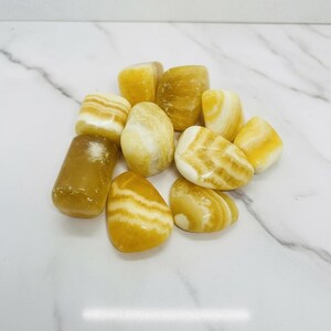 Puede incluir: Una colección de piedras de calcita amarilla pulidas con bandas blancas y amarillas. Las piedras varían en tamaño y forma, con superficies lisas y pulidas. Las piedras están dispuestas sobre una superficie de mármol blanco.