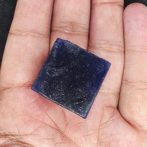 May include: A square, dark blue gemstone with a slightly rough texture. The gemstone is held in a hand, and the color is a deep, saturated blue. The edges of the stone are slightly irregular.
