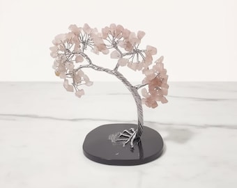 Pode incluir: Uma escultura decorativa de árvore com um tronco e galhos torcidos prateados. Os galhos são adornados com pedras de quartzo rosa claro, e a árvore assenta sobre uma base preta e redonda. A escultura é um item decorativo.