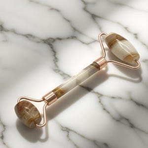 Puede incluir: Un rodillo facial con un mango y dos extremos redondeados. El rodillo está hecho de una piedra crema, marrón y beige, con detalles metálicos en oro rosa. El producto está diseñado para el cuidado de la piel y las rutinas de belleza.
