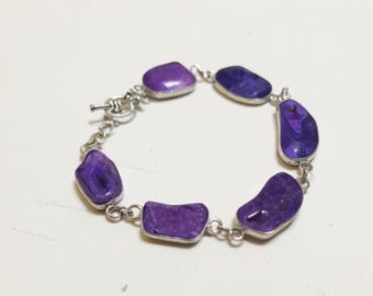 May include: A silver bracelet with seven polished, irregularly shaped purple stones. The stones are linked by a delicate chain and secured with a toggle clasp. The bracelet is displayed on a white surface.