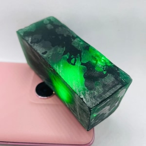 Könnte beinhalten: Ein rechteckiger Block aus rohem Jade, der einen leuchtenden Grünton mit dunkleren, gesprenkelten Mustern aufweist. Die Oberfläche des Steins ist rau und unpoliert und zeigt seine natürliche Textur. Der Jade liegt auf einer rosa Oberfläche.