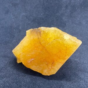 May include: A rough, golden-yellow mineral specimen with a crystalline structure. The stone has a slightly translucent quality, with variations in color intensity. The edges are irregular, and the overall shape is somewhat triangular. The mineral appears to be a natural, unpolished stone.