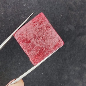 May include: A square, deep red object is held by silver tweezers against a dark background. The object has a textured surface and appears to be a sample of some kind. The tweezers are held by a person's hand.