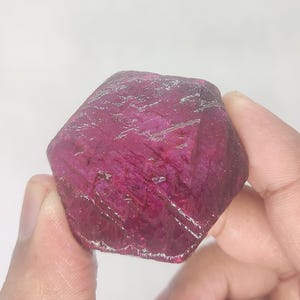 May include: A raw ruby gemstone with a deep magenta hue, held between fingers. The faceted surface displays a textured appearance with visible crystalline structures. The gemstone's natural form is highlighted against a white background.