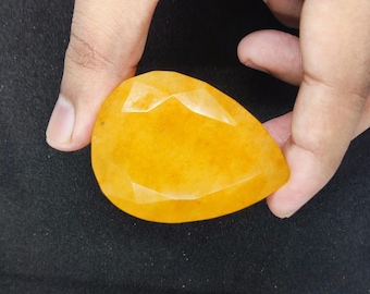 May include: A faceted, teardrop-shaped gemstone in a vibrant yellow-orange hue. The gem is held between two fingers, showcasing its polished surfaces and intricate cuts. The stone's color is reminiscent of a ripe mango.