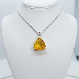Trillion yellow sapphire pendant necklace closeup inside white jewelry box, faceted golden gemstone with polished prongs and decorative bail, statement jewellery gift for women, uplifting sunshine color accessory associated with confidence and prosperity intentions in crystal traditions.