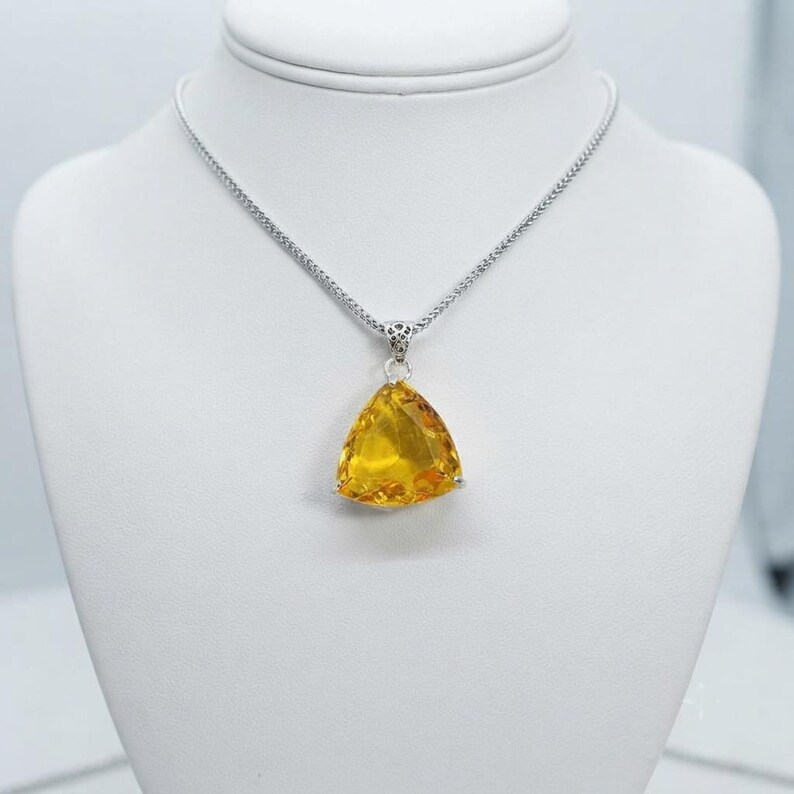 Trillion yellow sapphire pendant necklace closeup inside white jewelry box, faceted golden gemstone with polished prongs and decorative bail, statement jewellery gift for women, uplifting sunshine color accessory associated with confidence and prosperity intentions in crystal traditions.