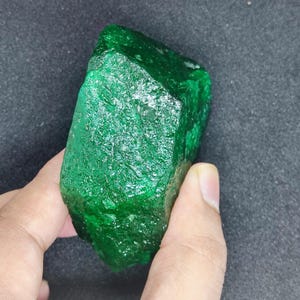 Può includere: Una gemma di smeraldo grezza e non tagliata. Lo smeraldo è di un colore verde intenso e vibrante, con una superficie ruvida e strutturata. La gemma è tenuta in mano su uno sfondo scuro.