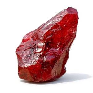 Puede incluir: Una gema de rubí en bruto sin cortar. El rubí es de color rojo intenso y saturado con una superficie rugosa y texturizada. La gema tiene forma irregular y parece translúcida.