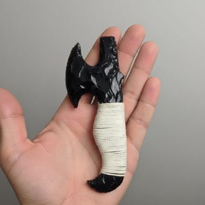 May include: A small, black obsidian axe with a curved blade and a wrapped handle. The handle is wrapped with white twine. The axe is held in a hand against a neutral background.