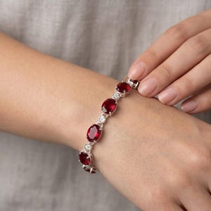 May include: A silver link bracelet featuring five oval-cut deep red gemstones. Each large red stone is separated by a small, circular silver setting containing tiny clear accent stones, creating a repeating pattern along the length of the bracelet.