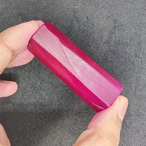 Peut inclure: Une pierre précieuse rectangulaire facettée de couleur magenta foncé. La pierre précieuse est tenue dans une main sur un fond gris foncé. La pierre a une surface polie et réfléchissante.