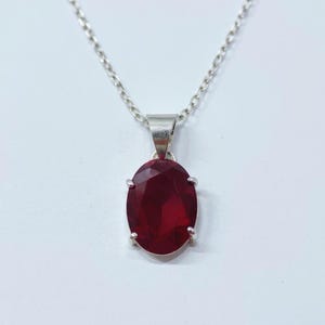 May include: A silver necklace featuring a large, oval-cut, deep red gemstone pendant. The pendant is set in a silver bezel and suspended from a delicate silver chain. The gemstone has a faceted surface, reflecting light.