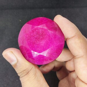 Pode incluir: Uma grande gema de rubi redonda e facetada. A gema é de uma cor magenta vibrante e é segurada entre dois dedos. As facetas criam um efeito brilhante, refletindo a luz na superfície. A pedra preciosa é um ponto focal, mostrando seu tamanho e cor.