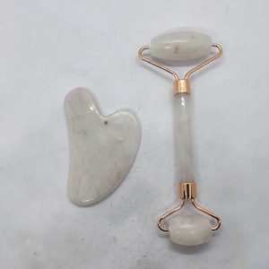 Op de afbeelding: Een gezichtsroller en een hartvormig gua sha-gereedschap. De roller heeft een witte steen en een roségouden metalen frame. De gua sha heeft een lichte, doorschijnende kleur. Beide tools zijn ontworpen voor huidverzorgingsroutines.