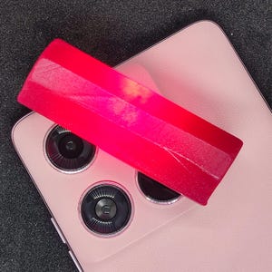 Könnte beinhalten: Ein leuchtend rosa rechteckiges Objekt mit strukturierter Oberfläche liegt auf einem rosa Smartphone. Das Telefon hat drei Kameralinsen. Der Hintergrund ist eine dunkelgraue Oberfläche.