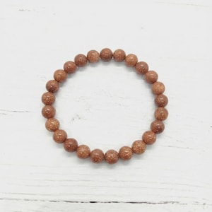 May include: A bracelet composed of reddish-brown, speckled beads. The beads are round and have a glittery appearance. The bracelet is displayed on a white, textured surface. Each bead is about 0.8 cm in diameter.