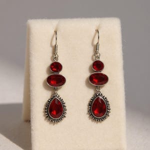 Puede incluir: Un par de pendientes colgantes plateados con tres piedras rojas. La piedra superior es redonda, la central ovalada y la inferior en forma de lágrima con un borde decorativo. Los pendientes se exhiben sobre una superficie blanca.